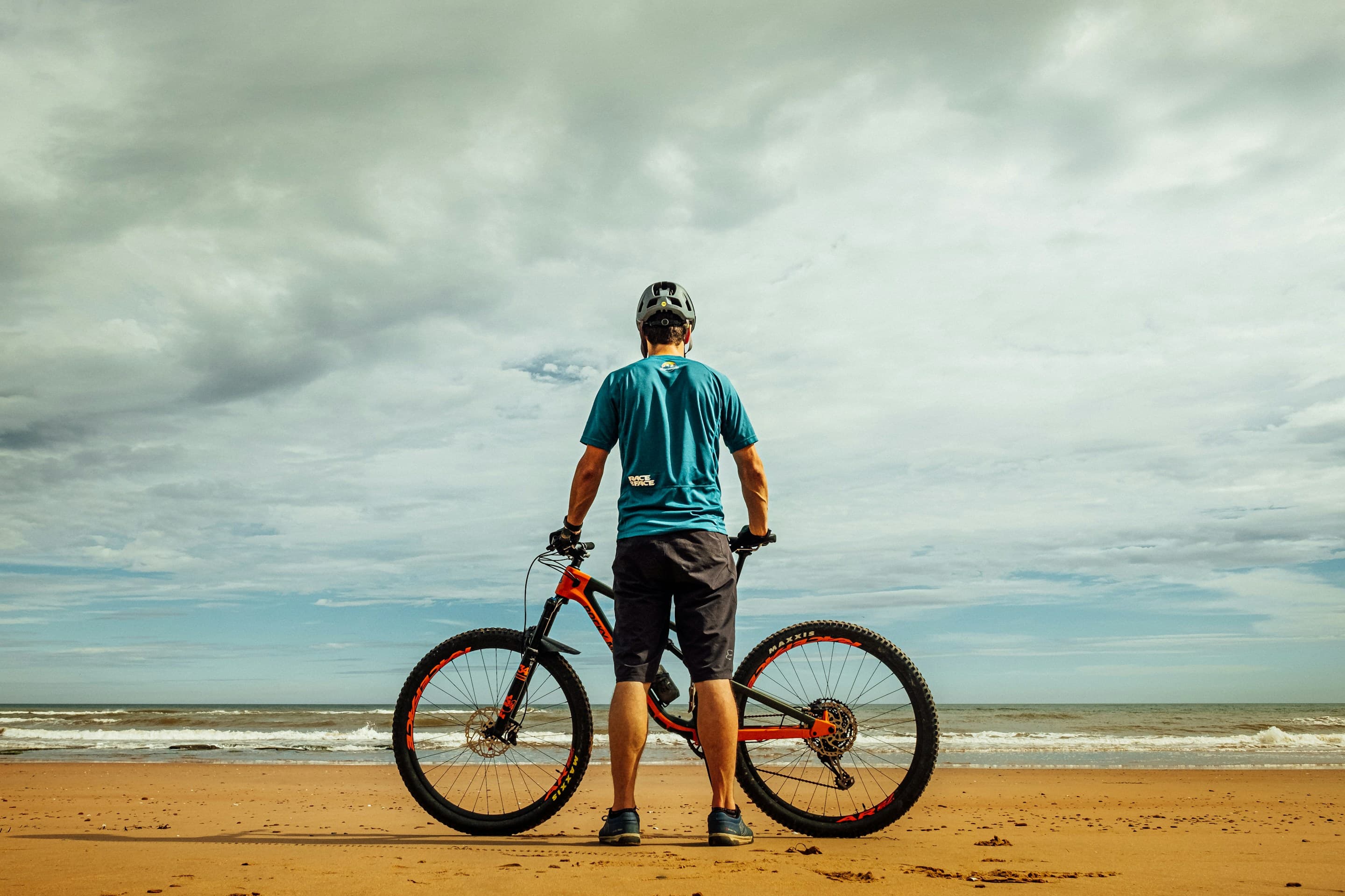A cyclist standing with their bike on the sand, looking out at the water from a beach